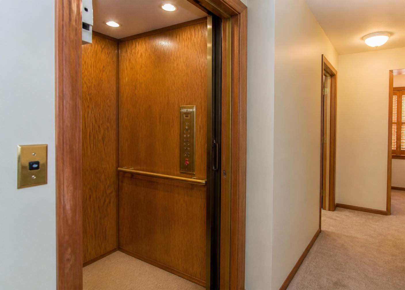 Oak-panelled residential elevator with a brass handrail opening onto a carpeted hallway