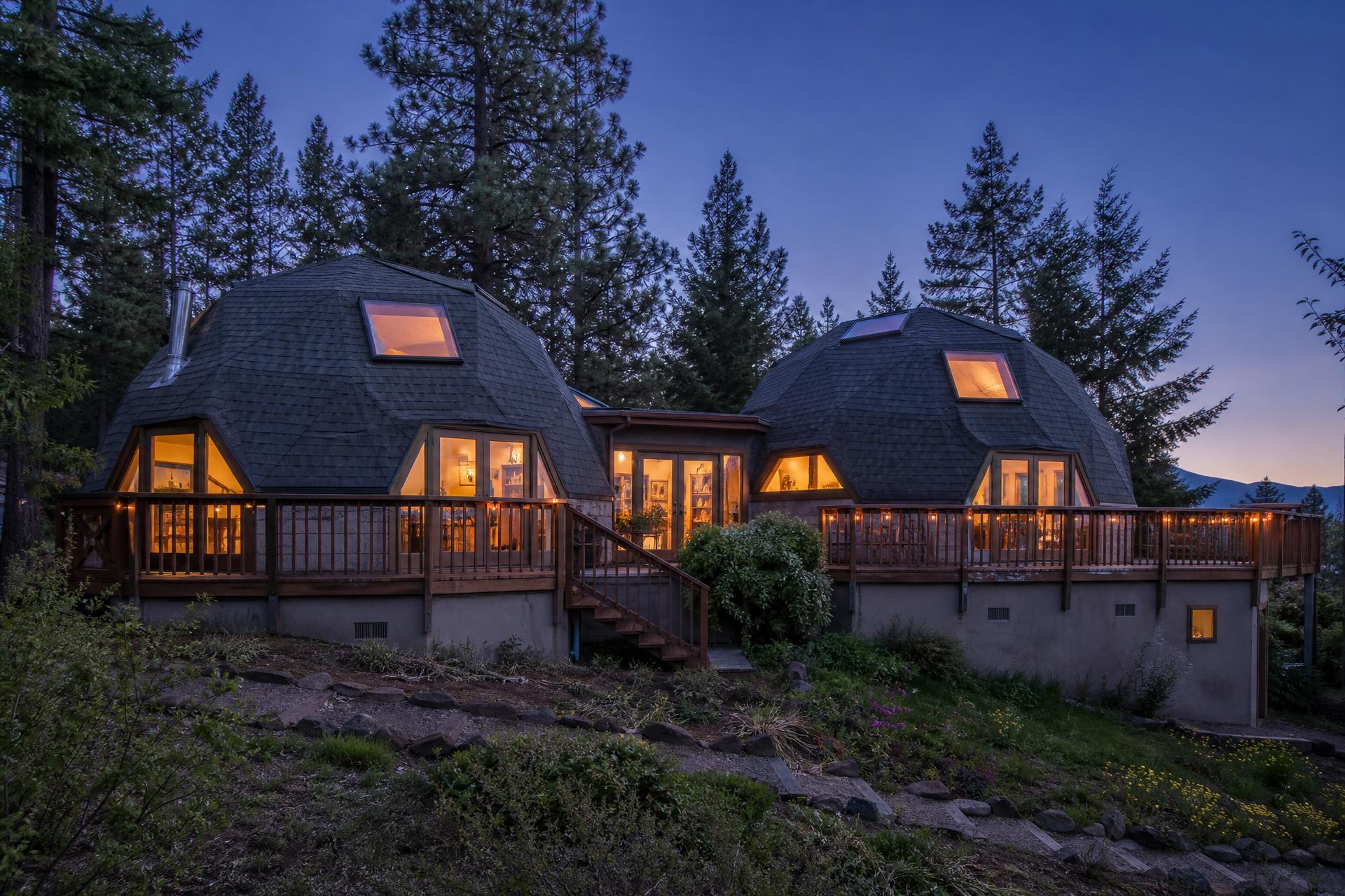 The double geodesic dome residence at dusk, on a traditional foundation with wrap-around deck and composition shingle roofing, warm light glowing through the windows