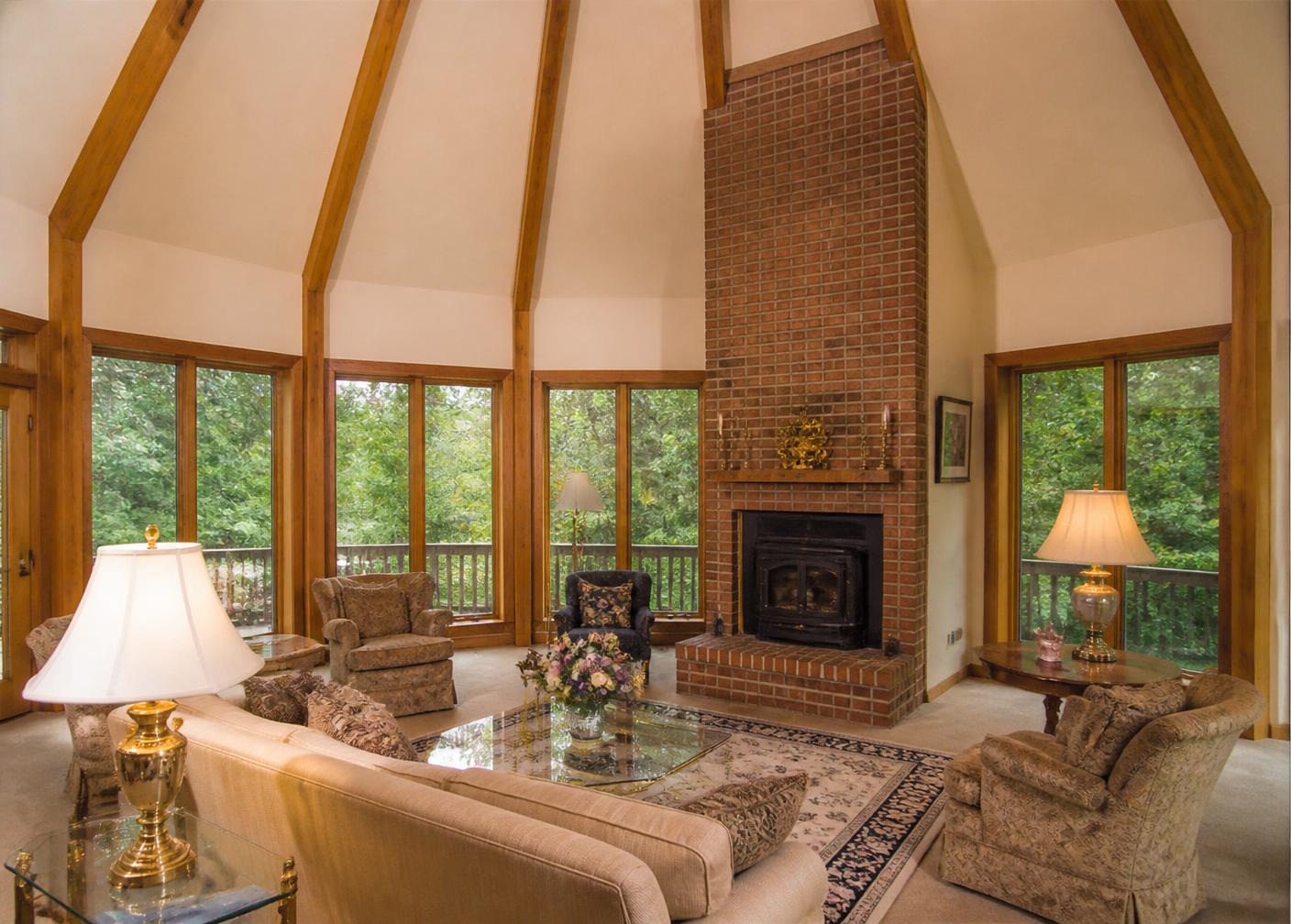 Great room with painted drywall ceiling planes, heavy oak beams, floor-to-ceiling brick fireplace, and oak-framed windows facing the forest